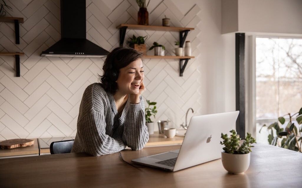 Young cheerful female smiling and talking via laptop while sitting at wooden table in cozy kitchen